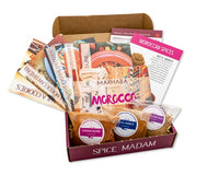 An open food subscription box labeled Spice Madam that is filled with 3 bags of spices and lots of recipe cards.