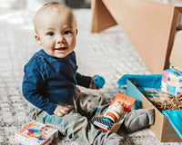 A baby boy sitting in front of a Hoppi subscription box and playing with baby toys from the box.