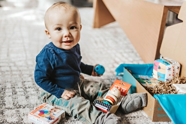 A baby boy sitting in front of a Hoppi subscription box and playing with baby toys from the box.