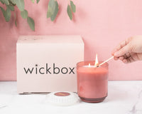 A hand using a match to light a candle in a pink jar, next to a closed Wickbox subscription box.